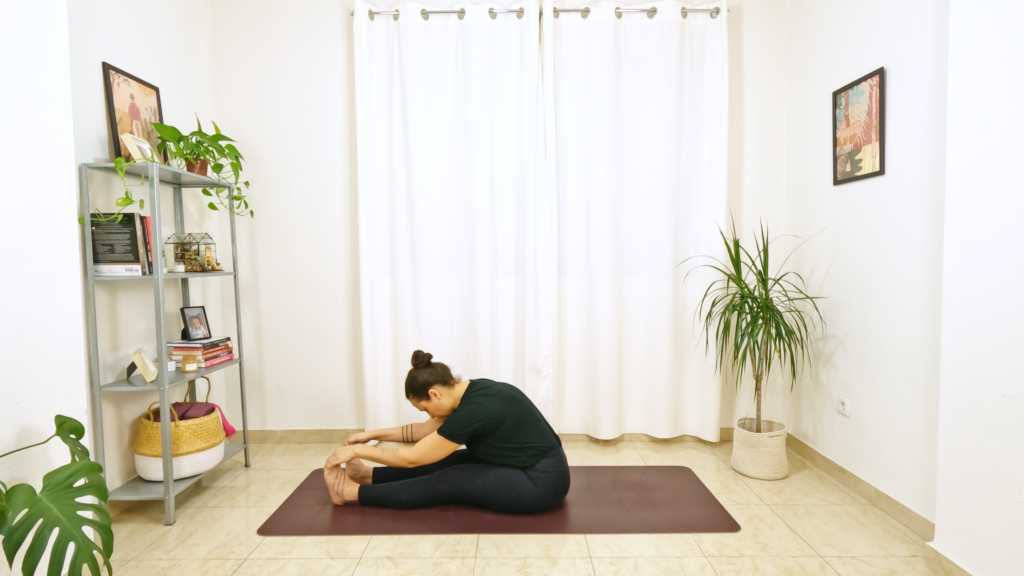 Mulher em postura de yoga sentada com as pernas esticadas e as mãos apoiadas nos pés