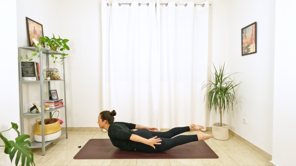 Mulher numa postura de yoga deitada de barriga para baixo com as pernas, peito e braços elevados, arqueando a coluna