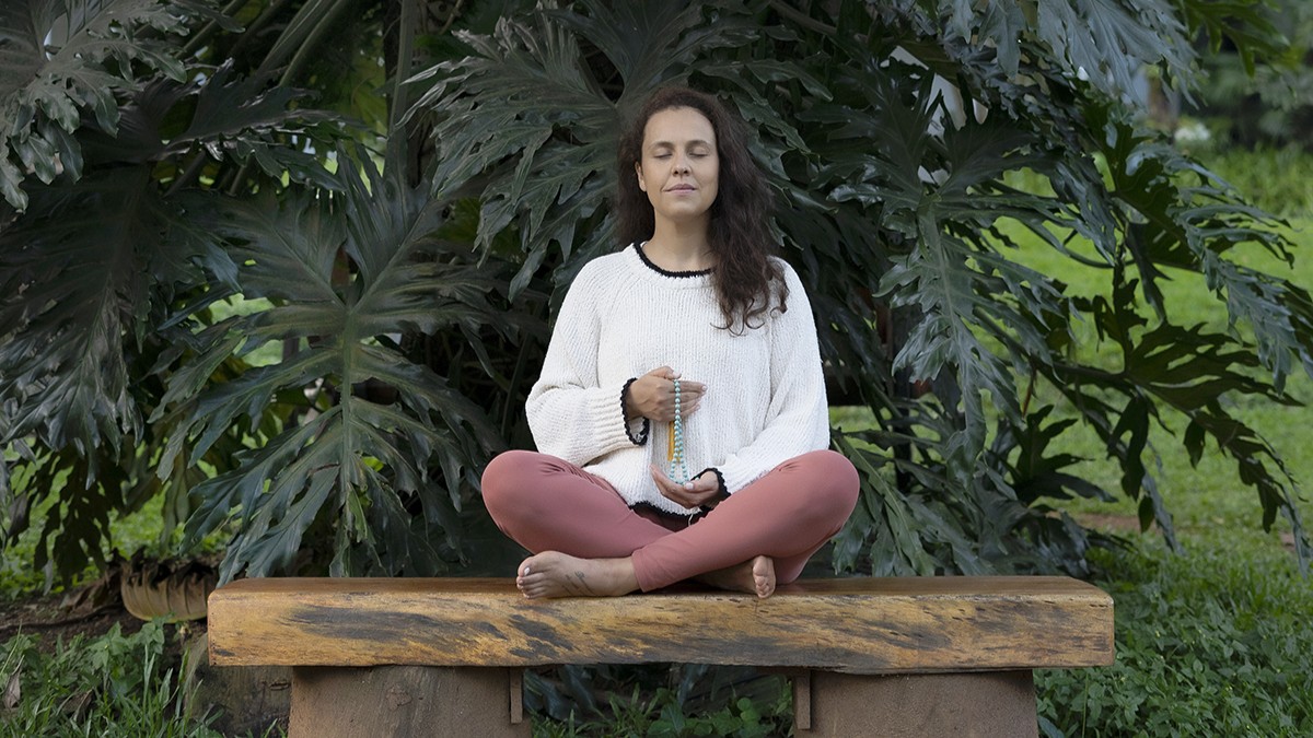 Mulher sentada em um bando de madeira em uma postura meditativa em frente a uma arvore.