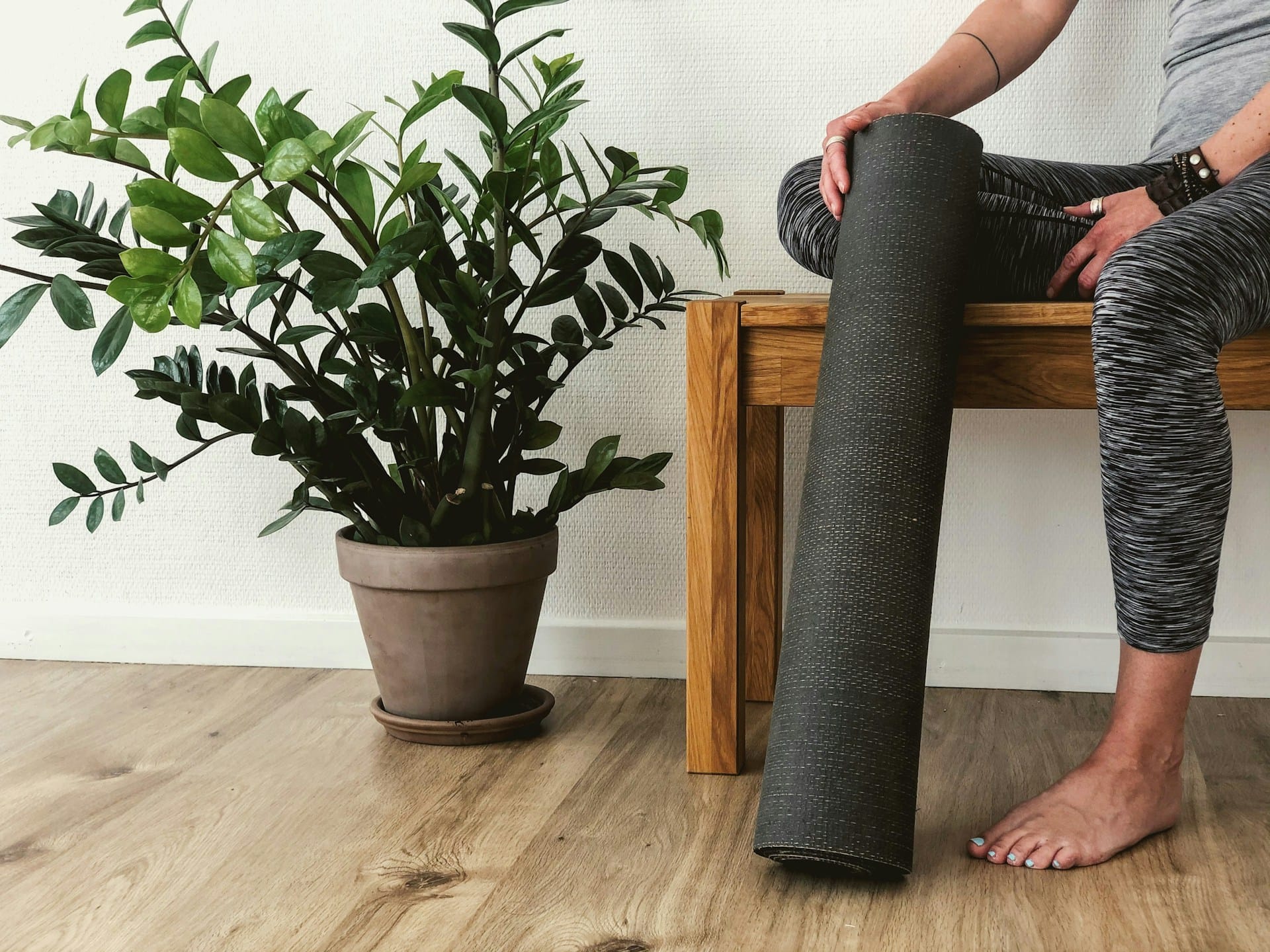 Foto de uma pessoa sentada em um banco de madeira segurando um tapete de yoga e ao lado de um vase de folhas verdes