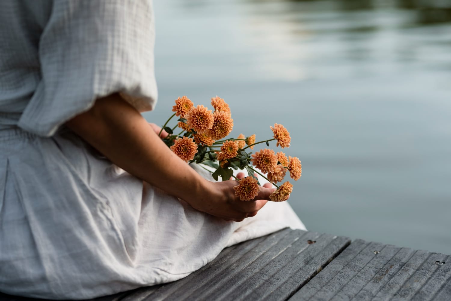 Mulher segurando um bique de flores laranjas sentada na frente de um corpo de agua